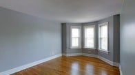 This single-floor space showcases a thoughtful blend of classic architectural character and clean, neutral aesthetics. The main living area is finished with warm-toned hardwood floors that add richness and depth to the room, complementing the soft gray walls painted throughout. A standout architectural feature is the bay window configuration in the corner of the room, where multiple double-hung windows with crisp white trim frame generous views of the surrounding greenery outside. This arrangement allows abundant natural light to flood the interior from multiple angles, enhancing the open and airy feel of the space. The windows feature traditional sill detailing, adding to the classic character of the room. White baseboards run along the perimeter, providing a clean contrast against the gray walls and tying together the overall aesthetic. The ceiling is smooth and finished in white, contributing to the sense of brightness and openness. The layout of the space appears versatile and well-proportioned, making it suitable for a variety of furniture arrangements. Overall, the space combines timeless architectural details with a neutral, modern palette.