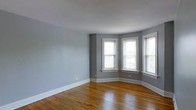 This single-floor space showcases a thoughtful blend of classic architectural character and clean, neutral aesthetics. The main living area is finished with warm-toned hardwood floors that add richness and depth to the room, complementing the soft gray walls painted throughout. A standout architectural feature is the bay window configuration in the corner of the room, where multiple double-hung windows with crisp white trim frame generous views of the surrounding greenery outside. This arrangement allows abundant natural light to flood the interior from multiple angles, enhancing the open and airy feel of the space. The windows feature traditional sill detailing, adding to the classic character of the room. White baseboards run along the perimeter, providing a clean contrast against the gray walls and tying together the overall aesthetic. The ceiling is smooth and finished in white, contributing to the sense of brightness and openness. The layout of the space appears versatile and well-proportioned, making it suitable for a variety of furniture arrangements. Overall, the space combines timeless architectural details with a neutral, modern palette.