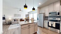 This thoughtfully designed one-bedroom, one-bathroom residence offers a cohesive blend of modern style and functional living across a single floor. The open-concept kitchen and living area serves as the heart of the home, where warm wood-look plank flooring flows seamlessly throughout. The kitchen is outfitted with crisp white cabinetry, granite countertops, a subway tile backsplash, and a full suite of stainless steel appliances including a French door refrigerator, electric range, microwave, and dishwasher. A center island with seating for two anchors the space, illuminated by a pair of striking black pendant lights with gold interiors. The adjoining living area features soaring ceilings, abundant natural light from large windows, and a ceiling fan overhead, creating an airy and inviting atmosphere. The bedroom is carpeted for comfort, features vaulted ceilings, a ceiling fan, and a large window, and provides access to a dedicated walk-in closet with built-in shelving and hanging rods. The full bathroom is finished with large-format tile flooring, a tiled tub-shower combination, a granite-topped vanity, and clean, neutral tones throughout. A dedicated entryway completes this well-appointed residence.