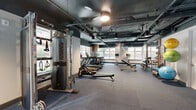 Located at 1031 N Vermont St in Arlington, VA, this fitness center offers a spacious, open-plan layout designed to accommodate a comprehensive range of workout needs. The space features an industrial-modern aesthetic, with an exposed ceiling showcasing dark-painted ductwork and structural elements, pendant lighting fixtures, and large windows along multiple walls that allow natural light to fill the room and provide views of the surrounding neighborhood.

The flooring throughout consists of durable carpet tile in a neutral gray tone, providing cushioning and traction suitable for a variety of exercise activities. Cardio equipment, including treadmills, ellipticals, and stationary bikes, is arranged along the windowed perimeter, taking full advantage of the natural light. A dedicated strength training area features a multi-station cable machine and functional training rig with suspension straps, while a well-stocked dumbbell rack provides a wide range of free weight options. A ball rack holds medicine balls, stability balls, and resistance tools for functional and flexibility training.

Warm wood accent columns punctuate the space, adding a touch of natural texture against the cool industrial palette. The entryway area includes a glass door with exterior access, abstract wall art, and a clean transitional zone. The overall ambiance is modern, energetic, and purposefully designed to support diverse fitness routines.