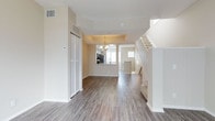This three-storey residence offers a well-organized layout designed for comfortable living across multiple levels. The main floor showcases an open-concept design, where the entryway flows seamlessly into a spacious living area finished with wide-plank, hardwood-style flooring in a warm grey-brown tone. Neutral beige walls and a textured ceiling contribute to a clean, understated aesthetic throughout.

A defining feature of the main level is the kitchen, which is partially open to the living space via a raised pass-through counter, allowing for easy interaction between the two areas. The kitchen is equipped with dark stainless steel appliances and white cabinetry, and is accented by a elegant chandelier overhead. A tray ceiling detail above the kitchen area adds architectural interest and a subtle sense of elevation.

A closet with louvred doors near the entry provides convenient storage, while a white-railed staircase with carpeted treads leads to the upper floors. Natural light filters through windows visible in both the kitchen and adjoining areas, keeping the space feeling bright and airy. The overall ambiance is modern, tidy, and move-in ready.