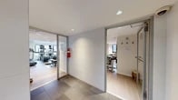Located at 26-38 21st Street in Astoria, New York, this combined lounge and gym amenity space offers a well-appointed, single-floor layout designed to serve both fitness and social needs. The space is divided into distinct functional areas connected by a central entry corridor, finished with glossy white tile walls, recessed lighting, and large-format floor tiles.

The fitness center is a standout feature, offering a generously sized, open workout floor with light wood flooring and bright, even lighting throughout. It is equipped with a full row of treadmills, elliptical machines, and other cardio equipment, alongside a dumbbell rack, weight benches, exercise mats, stability balls, and kettlebells. Floor-to-ceiling mirrors line one wall, amplifying the sense of space, while windows provide natural light and views of the surrounding neighborhood.

The resident lounge area provides a welcoming retreat with light hardwood floors, soft neutral tones, and stylish furnishings including a leather sofa, swivel accent chairs, and a high-top bar table with stools. A striking triptych canvas print of the New York City skyline serves as a focal point, reinforcing the urban character of the space. Shelving with books and board games adds a casual, communal feel.

A compact kitchenette serves the lounge, featuring dark countertops, a subway tile backsplash, a beverage cooler, and a sink — ideal for resident gatherings.