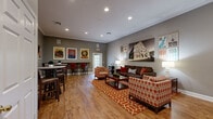 Located at 100 Riverchase Blvd in Nashville, TN, this community lounge is a well-appointed gathering space that celebrates its Music City roots through thoughtful design and décor. The single-floor layout is finished with warm hardwood-style flooring throughout, complemented by neutral gray walls and crisp crown molding that lend a polished, cohesive look.

The main seating area anchors the room with a rich brown sofa and a pair of boldly patterned armchairs in orange and ivory, arranged around a wooden coffee table atop a vibrant geometric area rug. Recessed ceiling lighting and freestanding floor lamps provide layered illumination across the space.

A dedicated media wall features a wall-mounted flat-screen television flanked by gray storage cabinetry with a warm wood countertop, accented by potted topiaries. An adjacent bar-height counter with red upholstered stools offers additional seating, ideal for casual gatherings or individual use.

Nashville-themed framed posters and artwork — including iconic imagery of the Ryman Auditorium, Music City trolleys, and local music culture — are displayed prominently throughout, giving the space a distinctive local character. A glazed exterior door allows natural light to filter in, brightening the room. Additional seating and a secondary TV area provide versatile functionality, making this lounge well-suited for both social and leisure activities.