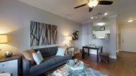 This single-floor residence showcases a thoughtfully designed open-concept layout that seamlessly connects the living, dining, and kitchen areas. The living room serves as the centerpiece, featuring rich hardwood flooring, elegant crown molding, and a ceiling fan that adds both style and functionality. A large, bold abstract artwork anchors one wall, complementing the neutral gray tones that define the space. The room accommodates a tufted gray sectional sofa accented with decorative pillows, paired with a glass-top coffee table, creating a comfortable yet polished atmosphere. Track lighting and a table lamp provide layered illumination throughout.

Adjacent to the living area, the dining space offers a sleek rectangular table set for entertaining, positioned to maintain an open flow between rooms. The kitchen, visible through a pass-through opening, features white cabinetry and appears well-equipped for everyday use. The overall design aesthetic is contemporary and cohesive, with a cool gray palette, warm wood flooring, and carefully curated décor that give the space a modern, inviting character throughout.