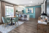 This single-floor leasing office combines professional functionality with a warm, inviting aesthetic. The main reception area serves as the centerpiece of the space, featuring a substantial walnut-toned L-shaped desk with seating for visitors, recessed lighting, and crown molding detail. Hardwood-style flooring runs throughout, complemented by patterned area rugs and coordinated drapery. A signature teal accent wall anchors the reception zone, adorned with contemporary abstract artwork and a round decorative mirror, lending the space a cohesive, modern mid-century character. A comfortable seating nook with teal upholstered chairs and framed wall art provides a welcoming area for guests. Natural light filters in through multiple windows dressed with patterned curtains, offering views of the surrounding outdoor areas. The kitchenette is well-equipped with white cabinetry, a granite-topped peninsula with bar seating, a sink, and a teal accent wall featuring bold artwork. An adjacent dining nook includes a round wood table with teal chairs and a large window overlooking an outdoor patio space, creating a bright and pleasant atmosphere. The space is unified by a consistent palette of teal, warm wood tones, and neutral whites, resulting in a professional yet approachable environment throughout.