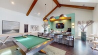 Located at 11107 Conductor Cir in Greer, South Carolina, this single-story clubhouse offers a thoughtfully designed communal space characterized by striking architectural details and versatile gathering areas. The vaulted ceiling with exposed wood beams serves as a defining architectural feature throughout, lending the interior a sense of openness and warmth.

The main lounge area is expansive and well-lit, anchored by a large sectional sofa, decorative coffee tables, and a wall-mounted flat-screen TV. A chandelier and recessed lighting complement the natural light admitted through tall French doors, which open to views of the surrounding community. Dark hardwood-style flooring flows throughout, providing a cohesive and polished aesthetic.

Adjacent to the lounge, a dedicated recreation area features a full-size billiards table positioned beneath statement pendant lights with copper interiors. A bar and counter seating area nearby is highlighted by a mint green shiplap accent wall, built-in cabinetry, a coffee station, and a granite-topped island with modern bar stools.

A bright café-style nook near the French doors offers a round bistro table with seating and abundant natural light, making it an inviting spot for casual conversation. The overall ambiance is contemporary yet welcoming, blending refined design elements with functional, community-oriented spaces.