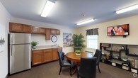 This family room is a thoughtfully designed communal space intended to provide a warm and comfortable environment for gatherings. The room features a functional kitchenette along one wall, complete with warm wood cabinetry, a countertop with a sink, and ample storage — ideal for preparing light refreshments during visits. Bright overhead fluorescent lighting ensures the space is well-lit throughout.

At the center of the room, a round wooden dining table is surrounded by padded black chairs, creating an inviting setting for shared meals or conversation. A window fitted with dark curtain valances allows natural light to filter in while maintaining a sense of privacy. A wall-mounted decorative cycling poster adds a touch of personality and visual interest to the space.

Lush potted plants placed around the room bring a refreshing touch of greenery, contributing to the overall welcoming ambiance. Carpeted flooring adds warmth underfoot, while a small shelving unit along one wall provides additional storage and display space. The neutral color palette of soft yellows and whites keeps the atmosphere bright and inviting, making this family room a comfortable retreat for meaningful time spent together.