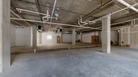 Located at 2323 Dulles Station Blvd in Herndon, Virginia, this single-floor commercial shell space presents a versatile, open canvas suited for a wide range of business uses. The space is currently in raw shell condition, offering tenants the opportunity to customize the interior entirely to their specifications.

The structure is built with exposed concrete columns, ceilings, and polished concrete slab flooring throughout, lending an industrial character to the space. Overhead, an extensive network of white-painted plumbing and mechanical pipes runs across the exposed concrete ceiling, with electrical panels and utility infrastructure already in place to support future build-out.

The space is divided into multiple open zones, including interior areas with open floor plans supported by concrete columns, and a front-facing section featuring large glass windows and glass double-entry doors that provide direct street access and abundant natural light. This storefront area offers strong visibility and a welcoming entry point.

Additional enclosed rooms and partially framed partitions are visible in sections of the space, indicating infrastructure for future subdivision. A gravel-lined utility trench runs through one interior area, suggesting provisions for plumbing installation. Overall, the space offers a functional, flexible foundation for commercial, retail, or service-oriented tenants.