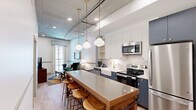 Located at 808 Constance St in New Orleans' Lower Garden District, this thoughtfully designed 2-bedroom, 1-bathroom space blends industrial character with refined modern finishes. Light wood flooring flows throughout, complementing exposed concrete ceilings and crisp white walls that give the interior a loft-inspired aesthetic.

The open kitchen and living area forms the heart of the space. The kitchen features two-tone cabinetry — navy blue lower cabinets and white uppers — paired with a farmhouse sink, gold brass hardware, subway tile backsplash, and a full suite of stainless steel appliances. A long wooden island with bar stools anchors the space and provides additional dining and prep area. Schoolhouse pendant lights hang overhead, adding warm character. The living area at the end of the corridor features a teal-painted ceiling, large windows with natural light, and an intimate seating arrangement.

The primary bedroom includes elevated ceilings with exposed concrete detail, a walk-in closet, wall-mounted TV, and a large window. Both bedrooms offer a calm, well-scaled retreat. The bathroom is elegantly appointed with a navy vanity featuring cane cabinet fronts, gold fixtures, a framed mirror with brass sconce lighting, and a glass-enclosed tiled shower. In-unit stacked laundry completes this efficiently designed space.
