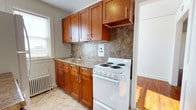 Located at 910 New Holland Ave in Lancaster, Pennsylvania, this single-floor space offers a practical and well-appointed layout designed for comfortable everyday living. The kitchen is a standout feature, showcasing rich, warm-toned wood cabinetry that extends floor to ceiling, maximizing storage capacity. Granite-style countertops provide a durable and attractive work surface, complemented by a textured stone tile backsplash that adds depth and visual interest to the space. A white electric coil range and a stainless sink are neatly integrated into the layout, offering all the essentials for a fully functional cooking space. Natural light filters in through a window adjacent to the sink, brightening the room and creating an open, airy feel. A classic cast-iron radiator is visible along the wall, indicative of a traditional heating system that provides consistent warmth. Just beyond the kitchen, warm hardwood flooring transitions into the adjacent living area, hinting at the cohesive and welcoming character that carries throughout the rest of the space. The overall design balances classic architectural elements with updated finishes, resulting in a space that is both timeless and practical for daily use.