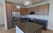 This 760-square-foot, one-bedroom, one-bathroom apartment at 2380 Staples Rd in San Marcos, TX, presents a clean, modern aesthetic throughout its single-floor layout. Light wood-tone flooring runs seamlessly across all living areas, complementing crisp white walls and bold matte black hardware accents. The open-concept kitchen and living room serves as the heart of the unit, featuring a striking kitchen with warm wood upper cabinetry, dark lower cabinets, dark granite countertops, a central island with an undermount sink and pendant globe lighting, and stainless steel appliances including a French door refrigerator and dishwasher. The living area is bright and airy, with multiple windows fitted with horizontal blinds, recessed lighting, and a ceiling fan with direct access to a private balcony. The 150-square-foot primary bedroom continues the light flooring and features a ceiling fan, natural light from a window, and a dedicated closet. The bathroom is finished with a dark vanity, granite countertop, undermount sink, matte black fixtures, and a tub-shower combo with tile surround. A dedicated laundry room houses a full-size side-by-side washer and dryer with an overhead storage shelf, completing this well-appointed apartment.