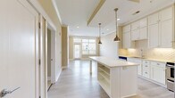 Located at 7511 Solstice Cir in Orlando, Florida, this 1-bedroom, 2-bathroom residence offers a thoughtfully designed single-floor layout with a bright, open-concept living space. The kitchen is a standout feature, showcasing white shaker cabinetry with glass-front uppers, a decorative arabesque tile backsplash, stainless steel appliances including a range with a sleek hood vent and a French door refrigerator, and a large center island with built-in shelving and a crisp white countertop. Antique brass pendant lights add a warm, stylish touch overhead. Light hardwood-style flooring flows throughout the main living and kitchen areas, while the bedroom features plush carpeting for added comfort. Large windows with blinds allow abundant natural light and offer views of the surrounding neighborhood. The primary bedroom is generously proportioned and connects to a well-appointed en-suite bathroom with a vanity, tile flooring, and a glass-enclosed shower, alongside a dedicated walk-in closet with wire shelving. The unit also includes a utility room, in-unit laundry, and access to a private balcony. Crown molding and a neutral warm-beige color palette give the space a clean, cohesive aesthetic throughout.