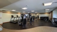 This fitness center offers a comprehensive workout environment designed to accommodate a range of exercise needs. The space is outfitted with multiple treadmills along one wall, providing ample options for cardiovascular training. A selection of cable and multi-use strength machines occupies the central area, offering versatility for resistance-based workouts. Along the far wall, a full dumbbell rack holds an organized array of free weights, catering to users of varying fitness levels. Large mirrors line the walls, enhancing the sense of openness while allowing users to monitor their form during exercise. The flooring transitions between dark rubber matting in the equipment areas, which provides durability and safety, and carpeted sections near the perimeter. Overhead, flush-mounted panel lighting ensures the entire space is evenly and brightly illuminated. The neutral color palette of whites and warm tones, combined with the clean layout, contributes to a focused and functional atmosphere. The room's generous proportions allow for comfortable movement between stations without congestion.