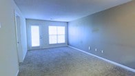 This single-story space presents a clean, functional living area finished in a neutral gray-blue palette that creates a calm, understated atmosphere. The main room is carpeted in a light, plush textile that adds warmth and comfort underfoot, while white baseboards provide a crisp contrast along the perimeter. Natural light enters generously through a large double-pane window fitted with horizontal blinds, allowing for adjustable brightness throughout the day. Adjacent to the window, a door with a built-in window panel offers an additional source of light and potential access to an outdoor area. The textured ceiling adds subtle visual interest overhead, and a centrally placed vent indicates a climate control system integrated into the space. Multiple electrical outlets are positioned along the walls, offering practical flexibility for furniture arrangement and electronics placement. The room's open layout provides versatile use of the available floor space, making it adaptable for a variety of living configurations. Overall, the space presents a tidy, move-in ready environment with a simple, neutral design well-suited for personalization.