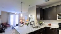 This single-floor residence showcases a thoughtfully designed open-concept layout that connects the kitchen and living area in a cohesive, functional flow. The kitchen is appointed with rich, dark cabinetry that provides a striking contrast against the light granite countertops, which offer generous prep and entertaining space. A stainless steel undermount sink sits beneath a sleek pull-down faucet, while two cylindrical pendant lights hang overhead, adding focused illumination and a contemporary decorative touch. The cabinetry features brushed nickel hardware, contributing to the modern aesthetic throughout. The kitchen transitions naturally into the living area, where neutral-toned walls and light flooring create a warm, welcoming ambiance. A ceiling fan enhances comfort, and large windows dressed with blinds and sheer curtains allow ample natural light to fill the space. The living area is comfortably proportioned, with enough room to accommodate a full seating arrangement. Together, the spaces offer a cohesive, modern interior defined by clean lines, quality finishes, and an open, airy feel suited to everyday living and entertaining.