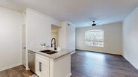 This well-appointed single-floor residence offers a thoughtfully designed open-concept layout that seamlessly connects the kitchen, dining, and living areas. Rich, dark hardwood-style flooring runs throughout the main living spaces, complementing the crisp white walls adorned with elegant crown molding.

The kitchen is a functional centerpiece, featuring white shaker-style cabinetry with matte black hardware, granite countertops, a peninsula with an undermount sink and a matte black pull-down faucet, and a full suite of stainless steel appliances including a refrigerator, range, microwave, and dishwasher. Track lighting illuminates the workspace, while built-in shelving near the entryway provides additional storage and display options.

The living area is bright and airy, anchored by a large arched window that fills the room with natural light and frames views of surrounding greenery. A ceiling fan with a light fixture enhances comfort and airflow. A secondary bedroom also benefits from natural light through a generous window, offering a calm and versatile space.

The bathroom includes a full tub-shower combination with white tile surround, matte black fixtures, and hardwood-style flooring. The overall aesthetic is clean and modern, with a neutral palette that creates a welcoming and cohesive atmosphere throughout.