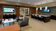This thoughtfully designed single-floor space exudes a polished, contemporary aesthetic that blends comfort with sophistication. The open-concept layout seamlessly connects a relaxed lounge area with a sleek bar and dining section, creating a versatile environment suited for both casual gatherings and more structured social settings.

The lounge area is anchored by a pair of upholstered gray armchairs accented with warm orange throw pillows, arranged around a clean white side table. Rich wood-paneled walls serve as a striking backdrop, adding warmth and texture to the space. A wall-mounted flat-screen television enhances the area's functionality, while recessed ceiling lighting provides an even, inviting glow.

An adjacent room, visible through large sliding wood-paneled doors, offers additional seating and dining options, expanding the overall capacity of the space. The bar counter area features elegant granite or stone-topped counters with high-backed white upholstered stools, creating a refined contrast between light and dark tones. Stylish pendant lights hang above the bar, adding a modern design focal point.

Light wood-toned flooring runs throughout, unifying the various zones while complementing the neutral color palette. Thoughtful decorative touches, including fresh floral arrangements and curated wall art, complete the welcoming and well-appointed atmosphere of this versatile communal space.