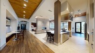 This expansive single-story clubhouse is designed with an open-concept layout that seamlessly connects multiple functional zones, creating a refined yet welcoming communal environment. The architectural character is defined by soaring vaulted ceilings clad in warm natural wood planking, contrasted against crisp white walls and large black-framed floor-to-ceiling windows that flood the interior with abundant natural light and frame views of the outdoor pool and surrounding grounds.

The lounge area serves as a central gathering point, anchored by a dramatic floor-to-ceiling marble fireplace wall with a built-in linear hearth and a wall-mounted flat-screen television. Seating includes tufted leather chairs, a tailored sofa, and accent chairs arranged around coffee tables, complemented by decorative lattice wood wall panels and curated artwork. An adjacent sitting nook features additional tufted leather seating and a wall-mounted display screen set within a textured recessed niche.

The kitchen and dining zone is equipped with stainless steel appliances, a gas cooktop beneath an oversized stainless range hood, bar-height seating at an island, and casual bistro-style tables positioned near expansive windows overlooking the pool courtyard. Flooring transitions between large-format light tile and rich wood-look planking, reinforcing the spatial distinctions throughout the clubhouse while maintaining a cohesive, upscale aesthetic.
