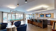 Located at 188 College St in New Haven, CT, this well-appointed amenity lounge offers a generously sized, open-plan layout spread across a single floor. The space is designed to encourage social interaction and relaxation, combining multiple distinct activity zones within one cohesive environment.

The lounge area anchors the space with a plush sectional sofa set against a bold yellow accent wall, complemented by a large wall-mounted television, a floating wood media console, and a curated gallery of framed artwork. Rich tones of gray and gold bring a vibrant, contemporary energy to this seating zone.

At the heart of the space, a substantial bar counter with a dark stone top and warm wood base is lined with modern upholstered barstools and illuminated by a trio of amber pendant lights. Built-in cabinetry and an embedded television make it an ideal gathering hub.

The game area features both a billiards table and a shuffleboard table, bathed in natural light streaming through expansive floor-to-ceiling glass doors that open directly onto an outdoor patio. Light wood-look flooring flows throughout, unifying each zone, while a coffered ceiling with layered lighting adds architectural interest. The seamless indoor-outdoor connection and multifunctional layout make this lounge a standout communal amenity.