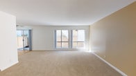 This single-floor space offers a clean, open layout designed for comfortable everyday living. The main living area is generously proportioned, featuring wall-to-wall carpet in a warm neutral tone that adds softness underfoot and complements the overall light color palette throughout. The walls are finished in a soft beige, creating a welcoming and understated ambiance that suits a wide range of interior styles.

Natural light is a defining feature of this space. A pair of large, grid-style windows lines the main living area wall, allowing daylight to pour in freely and brighten the interior throughout the day. These windows also provide views to the exterior, adding a sense of openness and connection to the outside.

Clean white ceilings and crisp white baseboards provide a polished contrast to the warm-toned walls and flooring, lending the space a tidy, well-maintained appearance. A sliding door is also visible along one wall, offering convenient access to an adjacent area or storage. The overall design is straightforward and functional, with a neutral palette that provides a versatile backdrop ready for personalization. The space presents a bright, move-in ready environment with an airy, uncluttered feel throughout.