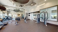 Located at 701 Little Palm Circle in Cape Coral, Florida, this well-appointed fitness center is designed to offer a comprehensive workout experience within a stylish, modern setting. The main exercise area features durable speckled rubber flooring and soaring ceilings with striking tiered tray and coffered details, ceiling fans, and recessed lighting that create an open, energizing atmosphere. Large windows and glass doors invite natural light and provide views of the surrounding Florida landscape, including glimpses of palm trees and a resort-style pool area beyond.

The facility is thoroughly equipped with a row of treadmills and elliptical machines for cardio training, a full dumbbell rack with adjustable benches, and a cable resistance machine for versatile strength work. Wall-to-wall mirrors amplify the sense of space and allow exercisers to monitor their form. A water fountain station and a neatly organized storage counter with rolled mats and accessories support a polished, user-friendly environment. Vibrant artwork adds a creative accent to the functional space.

A separate spin and cycling studio features warm hardwood flooring, mirrored walls, a mounted flat-screen TV, and a rack of medicine balls and resistance equipment, making it ideal for group or individual cycling and cross-training sessions. Throughout, the design balances modern aesthetics with practical functionality.