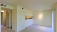 This single-floor residence on Breckinridge Court offers a clean, minimalist layout defined by bright white walls and neutral carpet flooring. The living area is open and unobstructed, creating a spacious feel enhanced by recessed track lighting that casts a warm glow across the room. A standout architectural detail is the built-in pass-through window that connects the kitchen to the living space, providing both functional convenience and an open, communicative flow between the two areas. The kitchen, visible through the pass-through, features a sink and counter space neatly tucked behind the dividing half-wall, keeping the cooking area distinct while maintaining visual connectivity. The ceiling-mounted HVAC vent ensures climate control is seamlessly integrated into the design without disrupting the clean aesthetic. Outlet placements are thoughtfully distributed along the baseboards, supporting practical everyday use. The overall ambiance is light, airy, and understated, offering a neutral canvas well-suited for a variety of interior styles and furnishing preferences.