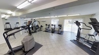 This fitness center is a thoughtfully designed single-floor exercise space that offers a comprehensive range of workout equipment within a clean, functional environment. The room features light-toned ceramic tile flooring throughout, providing a bright and hygienic surface that is easy to maintain. Rubber floor mats are strategically placed beneath the equipment to protect both the flooring and the machinery.

The space is equipped with a variety of cardio machines, including stationary upright and recumbent bikes, a stair climber, and additional aerobic equipment positioned along the perimeter. A multi-station cable weight machine anchors the strength-training area, offering users a versatile option for resistance workouts.

Large vertical windows with white vertical blinds line one wall, allowing generous amounts of natural light to fill the room while offering views of the outdoors. A wall-mounted air conditioning unit ensures a comfortable temperature during workouts. Overhead fluorescent lighting supplements the natural light, keeping the space well-illuminated at all times. The neutral white walls and open layout contribute to an airy, spacious atmosphere that supports a focused and comfortable exercise experience.
