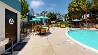 Located at 7400 Parkway Dr in La Mesa, California, this outdoor amenity area is managed by Bridge Property Management and offers a well-appointed poolside experience designed for relaxation and leisure. The centerpiece of the space is a generously sized, kidney-shaped swimming pool with clear turquoise water, framed by a smooth concrete pool deck that provides ample room for lounging and socializing.

Surrounding the pool, a collection of vibrant yellow-and-green lounge chairs and chaise lounges are thoughtfully arranged to take advantage of the abundant Southern California sunshine. A dedicated seating area adjacent to the pool features a round table with matching chairs shaded by large teal market umbrellas, creating an inviting spot for outdoor dining or casual gatherings.

The space is enclosed by a black iron perimeter fence, providing both security and a clean aesthetic boundary. Lush tropical landscaping — including tall palm trees, mature shade trees, and potted plants — frames the area, lending a resort-like atmosphere. The surrounding multi-story residential buildings are visible in the background, offering architectural context to the community setting.

Practical amenities are also present, including a conveniently located restroom facility with a drinking fountain situated just steps from the pool deck, ensuring comfort for all who use the space. Under the bright blue California sky, this outdoor area serves as a vibrant communal hub.