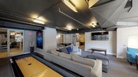 This single-floor amenity space is designed with a distinct industrial-modern aesthetic, characterized by dark, exposed ceilings with visible ductwork and piping, complemented by a combination of fluorescent and pendant lighting throughout. The space is thoughtfully divided into several functional zones that flow seamlessly into one another.

The lounge area serves as a central gathering point, anchored by a generously sized sectional sofa adorned with blue and gray accent pillows, positioned around a large-screen wall-mounted television. Adjacent to this is a dedicated billiards room, featuring a full-size pool table beneath a row of stylish pendant lights, with additional wall-mounted TVs and high-top tables with yellow-cushioned stools lining the perimeter.

A separate seating nook offers a more intimate setting, furnished with teal accent chairs and a coffee ottoman, positioned near large windows that frame outdoor views and allow natural light to filter in. Colorful artwork displayed on the walls adds personality throughout the space.

A dining area with a round table and upholstered chairs provides space for casual meals or gatherings. Carpeted flooring runs throughout, contributing to a comfortable, grounded atmosphere. The overall design balances urban industrial elements with inviting, well-curated furnishings.