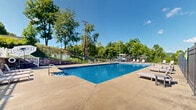 Located at 522 Blackstone Drive in Morgantown, West Virginia, this outdoor community amenity at Graycliff Place offers a well-appointed pool area designed for relaxation and recreation. The centerpiece is a generously sized rectangular swimming pool with crystal-clear blue water, accessible via built-in entry steps and handrail entries on multiple sides. Surrounding the pool is a broad concrete sundeck furnished with lounge chairs positioned to take full advantage of the open sky and natural sunlight. A shaded dining area with table seating and a patio umbrella provides a comfortable spot for outdoor gatherings. The pool area is fully enclosed by a black wrought-iron security fence, ensuring a safe and private environment. Stone masonry accents on the perimeter walls and stairways add a refined architectural touch. A covered deck extends from the adjacent building, offering additional outdoor space with views overlooking the pool. Lush mature trees and rolling green hills frame the backdrop, creating a scenic and serene atmosphere. The community entrance is marked by a stone monument sign and gated access, reflecting a well-maintained residential setting throughout.