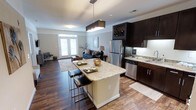 This single-floor residence showcases a thoughtfully designed open-concept layout that seamlessly connects the kitchen and living areas, creating a spacious and functional environment. The kitchen is a standout feature, highlighted by dark espresso cabinetry, granite countertops, and stainless steel appliances including a full-size refrigerator and dishwasher. A central island with a granite surface provides additional prep space and casual seating with modern bar stools, making it ideal for both cooking and entertaining. A built-in wine rack adds a sophisticated touch to the upper cabinetry. Rich, dark hardwood flooring extends throughout the open floor plan, lending warmth and elegance to the interior. The living area flows naturally from the kitchen, offering a comfortable and inviting atmosphere accented by neutral-toned walls. Natural light fills the space through a set of French doors and a transom window, brightening the interior and providing a welcoming connection to the outdoors. A modern rectangular pendant light fixture above the island serves as a contemporary focal point, complementing the overall aesthetic. The combination of dark cabinetry, granite surfaces, and hardwood flooring creates a cohesive, upscale ambiance throughout the home.