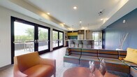 This thoughtfully designed single-floor community lounge space offers a dynamic and welcoming environment through its open-concept layout. The main lounge area features rich hardwood-style flooring, a tray ceiling with recessed lighting, and a bold teal accent wall adorned with framed abstract artwork. Colorful patterned sofas and leather accent chairs are arranged around a central oval coffee table, creating an inviting social atmosphere. A wall-mounted flat-screen television anchors the seating area, complemented by a striking arched floor lamp that adds a designer touch.

The kitchen area is a standout feature, boasting dark cabinetry with bold yellow and teal tile backsplash accents, stainless steel appliances, and a generous white-topped island with bar-height seating for multiple guests. Warm copper pendant lights hang above the island, adding an upscale, modern flair.

Large glass French doors line the exterior wall, flooding the space with abundant natural light and providing sweeping views of the surrounding greenery. These doors open directly onto a spacious outdoor deck furnished for additional seating and entertainment. The seamless transition between the interior lounge and the exterior deck makes this space well-suited for gatherings and social events.
