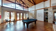 Located in Westworth Village, Texas, this single-story community clubhouse blends rustic charm with modern design in a spacious, welcoming environment. The interior is anchored by dramatic vaulted ceilings with exposed wood beam detailing and a striking antler chandelier that serves as a central focal point. Polished concrete floors in warm earth tones flow throughout, complementing the natural stone columns and a floor-to-ceiling stone fireplace fitted with an ornate iron screen and wall-mounted television above a wood mantel. The game room area features a full-size billiards table beneath the soaring ceiling, flanked by cue racks and bathed in natural light streaming through multiple sets of French doors fitted with sheer drapery. A comfortable lounge area provides seating with accent rugs, creating an inviting gathering space. A dedicated office or leasing area includes a substantial wood desk beneath the vaulted ceiling and chandelier, with additional storage cabinetry nearby. Throughout the space, French doors open to exterior views of the surrounding grounds, ensuring a strong connection to the outdoors. The combination of natural stone, warm wood tones, and abundant daylight gives the clubhouse a refined yet approachable Hill Country-inspired atmosphere.