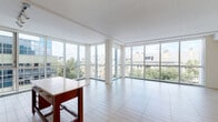 This single-floor residence is defined by its striking floor-to-ceiling glass walls, which wrap around the expansive open-plan living area and flood the space with natural light. Large-format light tile flooring runs throughout the main living areas, enhancing the bright, airy atmosphere, while panoramic views of the surrounding urban landscape create a dynamic backdrop. The living area is generously proportioned, with multiple window panels framing treetop and cityscape views from multiple angles.

The kitchen is thoughtfully designed with rich dark wood cabinetry featuring frosted glass upper panels, white quartz countertops, a pale green glass tile backsplash, and a full suite of stainless steel appliances. A portable island with built-in wine storage adds both function and warmth to the space.

The bedroom is carpeted for comfort and includes a large window with urban views, while the walk-in closet offers extensive wire shelving and hanging space. Two bathrooms serve the unit: one featuring a glass-enclosed walk-in shower, dual-sink vanity with dark wood cabinetry, and wood-look flooring; the other offering a streamlined single-sink vanity with a wide mirror. A tiled entry foyer provides a clean transition into the residence.