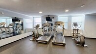 Located at 1317 East St in New Britain, CT, this dedicated fitness center offers a well-equipped exercise space designed to support a comprehensive workout routine. The room features durable speckled rubber flooring throughout, providing both safety and comfort during exercise. Recessed lighting illuminates the space evenly, complemented by natural light streaming in through multiple windows along the perimeter walls.

The cardio section is equipped with multiple treadmills and an elliptical trainer, all from recognized commercial-grade brands. A ceiling-mounted television is positioned for easy viewing during cardio sessions. For strength training, the space includes a multi-function cable machine, an adjustable weight bench, a rack of medicine balls, and a selection of dumbbells. An additional strength machine and a stretching mat offer further options for varied training.

Large wall-mounted mirrors enhance the sense of space and allow users to monitor their form during workouts. The walls are finished in a clean, neutral gray tone with white crown molding, giving the room a polished appearance. Subtle wall-mounted light fixtures and a textured ceiling complete the interior. The space is accessed through a solid gray entry door and includes sanitation stations for user convenience.
