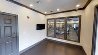 This single-floor fitness center is thoughtfully designed to support a comprehensive workout experience. The space is divided into distinct functional areas, offering both open exercise zones and a dedicated activity or stretching room. The activity room features a wall-mounted flat-screen television, recessed lighting, crown molding trim, and durable rubberized flooring — ideal for yoga, stretching, or low-impact workouts. Large glass-panel windows and doors connect this room to the main gym floor, maintaining an open and airy feel while providing visual connectivity throughout the space. The main workout area, visible through the glass partitions, is outfitted with an array of strength training machines, free weights, and cardio equipment including treadmills and ellipticals. Overhead ceiling fans and recessed lighting ensure the space remains well-lit and comfortable during workouts. The clean white walls, dark trim accents, and modern finishes lend the facility a polished, contemporary aesthetic. Overall, the fitness center is bright, functional, and well-maintained, making it a practical and inviting environment for a wide range of fitness activities.