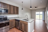 This single-floor residence is designed with an open-concept layout that seamlessly connects the kitchen and living areas, creating a spacious and functional flow. The kitchen is a standout feature, equipped with rich dark wood cabinetry, granite countertops, a white subway tile backsplash, and a full suite of stainless steel appliances, including a refrigerator, range, microwave, and dishwasher. A peninsula with an undermount sink extends into the living space, illuminated by a pair of modern pendant lights that add a contemporary touch. Warm hardwood-style flooring runs throughout the main living areas, complementing the neutral wall tones. The living area is fitted with a ceiling fan and receives natural light through a sliding glass door that opens to the outdoors. A separate room features a wide window with outdoor views of open green space, offering a bright and airy atmosphere. A bathroom with hardwood-style flooring is conveniently situated adjacent to a dedicated laundry area with a stacked washer and dryer unit. The overall ambiance of the space is clean, modern, and well-appointed, with consistent finishes tying each area together cohesively.