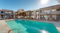 Located at 333 W Van Buren St in Avondale, Arizona, this outdoor amenity area serves as a central gathering space for the surrounding residential complex. The focal point is a generously sized swimming pool with clear turquoise water, enclosed by a white metal safety fence complete with posted safety signage and a life ring. The pool deck is finished in light-toned concrete and offers ample space for outdoor leisure. A dedicated sunbathing area features multiple chaise lounge chairs positioned to take full advantage of the Arizona sunshine. Adjacent to the pool, a shaded outdoor dining area provides a comfortable setting with a large patio umbrella, a dining table, and additional seating — ideal for socializing in the open air. Mature trees add natural shade and visual character to the courtyard setting. The surrounding two-story residential buildings display a Southwestern-influenced architectural style, with stucco exteriors, tile roof accents, and neutral earth-toned color palettes. Exterior staircases provide access between floors, and the buildings frame the pool area on multiple sides, creating an enclosed, community-oriented environment. Under the clear blue skies characteristic of the Avondale region, this outdoor space offers a well-appointed retreat designed for relaxation and community use.