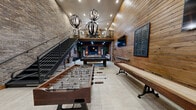 Located at 21409 Town Lakes Dr in Boca Raton, FL, this two-floor entertainment space delivers a striking industrial-rustic aesthetic across a generously sized layout. The main floor commands attention with soaring double-height ceilings that amplify the sense of openness, while exposed brick walls contrast beautifully with warm wood-paneled surfaces throughout. Large-format light tile flooring grounds the space with a clean, modern finish. The game area is a central highlight, featuring a foosball table, a shuffleboard table, and a pool table, all illuminated by dramatic globe pendant light fixtures suspended from above. A sleek steel staircase with cable railing rises along one wall, connecting the main floor to the upper level. Tucked beneath a rich wood-beamed ceiling, a cozy lounge area offers a plush sectional sofa, a wall-mounted flat-screen TV set against a deep teal accent wall, and warm recessed lighting that creates an intimate atmosphere. The upper floor, accessed via the staircase, provides an open mezzanine-style area overlooking the main floor below, framed by the same exposed brick walls and recessed lighting. Throughout, the layered use of natural materials, bold textures, and carefully curated lighting creates a cohesive and inviting environment ideal for recreation and entertainment.