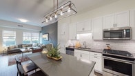Located at 100 N Addison Ave in Elmhurst, Illinois, this single-floor residence showcases a thoughtfully designed open-concept layout with modern finishes throughout. The kitchen serves as a focal point of the home, featuring crisp white shaker cabinetry, a classic white subway tile backsplash, dark quartz countertops, and a full suite of stainless steel appliances including a gas range, over-the-range microwave, dishwasher, and side-by-side refrigerator. A generously sized central island with bar seating provides both additional prep space and a casual dining option, illuminated by a striking industrial-style pendant light fixture. Rich hardwood flooring flows through the main living areas, connecting the kitchen seamlessly to the open living room. The living area is anchored by a deep-toned accent wall and large windows that invite abundant natural light and outdoor views. The bedroom offers plush carpeting, ample natural light through large windows, and a serene, neutral palette. The bathroom is finished with a white shaker vanity, dark quartz countertop, black fixtures, and hardwood-style flooring, maintaining a consistent modern aesthetic. The overall ambiance is contemporary and welcoming, blending clean architectural lines with warm, livable design elements throughout.