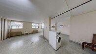 This single-floor communal laundry facility offers a generously sized, open layout designed for practical, shared use. The space is finished with durable speckled terrazzo flooring throughout, providing an easy-to-maintain surface well-suited to its function. Multiple windows along one wall face the surrounding neighbourhood, allowing natural light to fill the room while providing outdoor views and a sense of openness.

The laundry area is equipped with several front-loading and top-loading washing machines and dryers arranged for efficient use, accompanied by a utility sink and folding surfaces. Informational notices are posted on the walls, and a wall-mounted clock keeps the space functional and organized. Fluorescent ceiling fixtures supplement natural light throughout the day and evening.

A distinct section of the space features a bold striped wood-panel partition wall with integrated locker storage, adding both a visual design element and practical storage for residents. A stone-topped utility table is positioned nearby, offering additional workspace. Cast-iron radiators line the walls, indicating a traditional heating system. A solid entry door with secure hardware provides access to the room, ensuring a controlled and private communal environment.