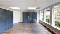 This package locker room is a well-appointed, single-floor space designed to provide a convenient and secure package retrieval experience. The centerpiece of the room is a bank of Amazon Hub smart lockers, spanning multiple units with a built-in touchscreen interface, offering residents a reliable and self-service solution for receiving deliveries of all sizes.

The space features warm wood-look flooring that runs throughout, complemented by neutral-toned walls and a drop ceiling with recessed panel lighting that keeps the room bright and welcoming. Large windows line the perimeter, flooding the interior with generous natural light and offering pleasant views of the surrounding community grounds, including manicured hedges, a water feature, and neighboring buildings.

Decorative touches such as an ornate console table, framed wall art, and potted plants add a polished, residential feel to what is primarily a functional space. A glass-panel entry door provides easy access, while an interior door connects to adjacent areas of the building.

Overall, the room strikes a balance between practicality and aesthetic care, ensuring that package pickup is not only efficient but also a comfortable experience within a thoughtfully designed environment.