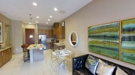 This thoughtfully designed single-floor residence showcases an open-concept layout that effortlessly unites the kitchen, dining, and living spaces into one cohesive, inviting environment. The kitchen is well-appointed with rich warm-toned cabinetry extending to the ceiling, offering generous storage, while stainless steel appliances — including a French door refrigerator and a built-in microwave — lend a modern, functional edge. A kitchen island with a stone countertop provides additional prep space and casual seating. Recessed lighting throughout the space ensures bright, even illumination, complemented by elegant pendant lights above the island. The dining area features a compact round table with sleek acrylic chairs, creating an airy and contemporary feel. A round wall mirror and a vibrant green abstract painting add personality and visual interest to the neutral beige walls. The living area anchors the space with a tufted black leather sofa accented with decorative pillows, offering a comfortable retreat. Large light-toned tile flooring flows continuously through the space, tying each area together and enhancing the sense of openness. The overall ambiance is modern, warm, and welcoming.