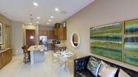 This thoughtfully designed single-floor residence showcases an open-concept layout that effortlessly unites the kitchen, dining, and living spaces into one cohesive, inviting environment. The kitchen is well-appointed with rich warm-toned cabinetry extending to the ceiling, offering generous storage, while stainless steel appliances — including a French door refrigerator and a built-in microwave — lend a modern, functional edge. A kitchen island with a stone countertop provides additional prep space and casual seating. Recessed lighting throughout the space ensures bright, even illumination, complemented by elegant pendant lights above the island. The dining area features a compact round table with sleek acrylic chairs, creating an airy and contemporary feel. A round wall mirror and a vibrant green abstract painting add personality and visual interest to the neutral beige walls. The living area anchors the space with a tufted black leather sofa accented with decorative pillows, offering a comfortable retreat. Large light-toned tile flooring flows continuously through the space, tying each area together and enhancing the sense of openness. The overall ambiance is modern, warm, and welcoming.