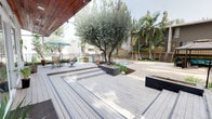 This inviting outdoor courtyard serves as a central communal space, thoughtfully designed to blend natural landscaping with functional amenities. The centerpiece is a generous multi-level composite deck finished in light grey tones, offering distinct zones for lounging, dining, and socializing. A partially covered section features a warm wood-paneled ceiling with recessed lighting, providing shade over a dining set complete with patio chairs and a teal umbrella — perfect for al fresco gatherings.

Mature trees, including tall pines, a prominent olive tree, and tropical palms, are integrated throughout the space, lending a lush, resort-like atmosphere. Raised dark planter boxes add structured greenery and define pathways across the deck levels. Potted plants and cultivated garden beds further enrich the natural ambiance.

A dedicated lounge area with a gazebo, cushioned seating, and a grill offers a comfortable retreat for relaxed outdoor entertaining. Beyond the deck, a pool area is visible, framed by iron fencing and additional landscaping, enhancing the overall sense of a well-appointed outdoor community space. Large sliding glass doors connect the interior to the deck, flooding the transition areas with natural light and maintaining a seamless indoor-outdoor flow throughout this single-level property.