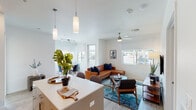 This well-appointed single-floor residence offers a thoughtfully designed open-concept layout that seamlessly connects the kitchen, dining, and living areas. The kitchen is outfitted with flat-front white cabinetry extending to the ceiling, quartz countertops, a white subway tile backsplash, stainless steel appliances including an electric range and over-the-range microwave, and a central island with pendant lighting overhead. The dining area is tucked beside large windows with roller shades, filling the space with natural light and offering views of the surrounding outdoors. The living room features light wood-style flooring, a ceiling fan, and multiple windows that bring in additional natural light, creating a bright and airy atmosphere. Two bedrooms provide comfortable retreats, each with ceiling fans, roller shade windows, and neutral carpeting. The bathroom is clean and functional, featuring a combined tub and shower with subway tile surround, a single vanity with under-sink storage, built-in shelving niches, and wood-style flooring. Throughout the residence, a consistent modern aesthetic is maintained with crisp white walls, brushed nickel fixtures, and an overall design sensibility that balances style with practicality.