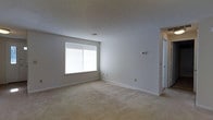 This single-story space presents a straightforward and functional layout with a clean, neutral aesthetic throughout. The main living area is finished with wall-to-wall carpet in a light beige tone, contributing to a warm and understated atmosphere. Smooth, light gray walls are complemented by white baseboards, creating a crisp contrast that keeps the space feeling bright and open. A sliding window allows natural light to fill the room, enhancing the airy quality of the interior. The textured popcorn ceiling adds a subtle dimension overhead, while a centrally located ceiling vent ensures efficient climate control. A wall-mounted thermostat is conveniently positioned for easy access. Electrical outlets are distributed along the walls, offering practical utility throughout. An adjoining hallway, visible from the main area, suggests additional rooms beyond, hinting at a connected and cohesive floor plan. Overall, the space offers a neutral, move-in-ready canvas well-suited for a variety of furnishing styles and functional uses.
