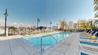 Located at 28 Southfield Avenue in Stamford, Connecticut, this outdoor recreation area offers a resort-style experience in a vibrant waterfront setting. The centerpiece of the space is a generously sized swimming pool, its crystal-clear blue water adorned with a signature anchor mosaic on the pool floor — a nod to the nautical character of the surrounding marina. The pool deck is expansive, providing ample room for rows of lounge chairs and side tables, ideal for sunbathing and relaxation throughout the warmer months. Classic-style lamp posts punctuate the deck, adding a refined aesthetic to the outdoor environment. Perimeter fencing frames the space while maintaining open sightlines to the marina, where sailboat masts rise against the skyline, creating a distinctive backdrop. A brick amenity building with a blue roof and bold orange entry doors anchors one end of the space, housing restroom and support facilities. Adjacent to the building, a turf lawn area provides a soft green contrast to the surrounding hardscape pavers. The entire area is finished with large-format stone tiles, lending a clean, contemporary feel. The surrounding multi-story residential buildings complete the enclosed courtyard atmosphere, making this a well-appointed and visually engaging outdoor amenity space.