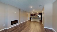 This single-floor unit presents a well-designed open-concept layout that seamlessly connects the living area and kitchen, creating a spacious and functional environment. Rich, dark hardwood flooring runs continuously throughout, complementing the warm neutral taupe wall tones and crisp white baseboards that define the interior palette.

The living area is anchored by a sleek, wall-mounted electric fireplace, providing a modern focal point. A textured ceiling adds subtle depth to the space, while recessed lighting ensures consistent illumination. A standard entry door with a sidelight window brings in a modest amount of natural light.

The kitchen is a standout feature, outfitted with deep espresso-toned shaker-style cabinetry extending to the ceiling for generous storage. Granite countertops in light grey tones span both the perimeter counters and a central island that doubles as a breakfast bar. Stainless steel appliances, including a refrigerator, range, microwave, and dishwasher, complete the kitchen. Pendant lighting above the island adds a refined touch. A set of double doors near the kitchen provides additional storage.

The bathroom mirrors the home's design language, featuring a dark vanity with granite countertop, a large mirror, and a combined tub-shower unit.
