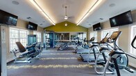 This single-floor fitness center is designed to support a comprehensive workout experience within a thoughtfully organized layout. The space is anchored by a striking vaulted ceiling with a ceiling fan and linear cove lighting that runs along the roofline, creating a sense of openness and volume. Accent walls painted in a bold olive green contrast with the neutral taupe and white tones throughout, lending visual energy to the environment.

The main workout area features a row of cardio equipment, including treadmills, upright and recumbent stationary bikes, and elliptical machines, all positioned to take advantage of natural light from multiple windows overlooking the outdoors. Large wall-mounted mirrors extend the perceived depth of the space while allowing users to monitor their form. Wall-mounted flat-screen TVs are strategically placed throughout for entertainment during workouts.

The strength training section includes a cable machine system, a dumbbell rack, and a weight bench, catering to a range of resistance training needs. A patterned carpet in charcoal and yellow tones covers the floor, with rubber mat zones beneath equipment for durability and safety.

Toward the rear, a dedicated area with built-in shelving, a cushioned bench, and a water station provides a practical space for storage and recovery. The layout flows intuitively from cardio to strength to cooldown zones.
